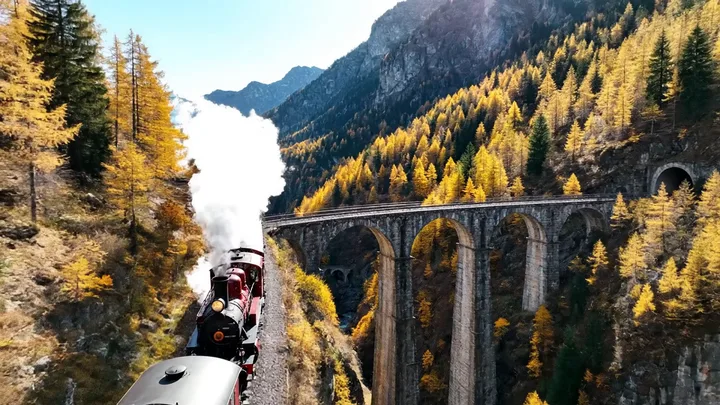 Aerial shot following a vintage red train across an alpine stone viaduct in autumn, generated with Veo 3.1 on FluxoKit